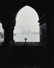 A silhouette of a camera on a tripod is framed by an archway, with a misty waterfront and distant buildings in the background. The scene is moody and atmospheric, with minimal visibility due to the fog.