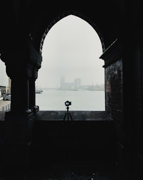 A silhouette of a camera on a tripod is framed by an archway, with a misty waterfront and distant buildings in the background. The scene is moody and atmospheric, with minimal visibility due to the fog.