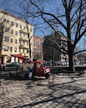 A red food truck is parked on a cobblestone street in an urban area, surrounded by tall buildings with European-style architecture. Leafless trees suggest a winter or early spring setting. Two folding chairs and a small table are placed in front of the truck, indicating a casual, outdoor dining setup.