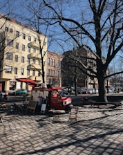 A red food truck is parked on a cobblestone street in an urban area, surrounded by tall buildings with European-style architecture. Leafless trees suggest a winter or early spring setting. Two folding chairs and a small table are placed in front of the truck, indicating a casual, outdoor dining setup.