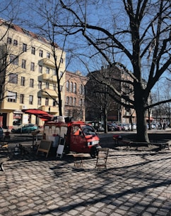 A red food truck is parked on a cobblestone street in an urban area, surrounded by tall buildings with European-style architecture. Leafless trees suggest a winter or early spring setting. Two folding chairs and a small table are placed in front of the truck, indicating a casual, outdoor dining setup.