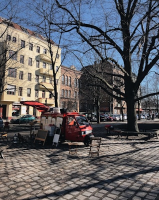 A red food truck is parked on a cobblestone street in an urban area, surrounded by tall buildings with European-style architecture. Leafless trees suggest a winter or early spring setting. Two folding chairs and a small table are placed in front of the truck, indicating a casual, outdoor dining setup.