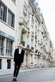 A person in a black suit and white shirt stands on an empty urban street with tall, elegant buildings lining the sidewalk. The architecture features classic European design, with balconies and large windows.