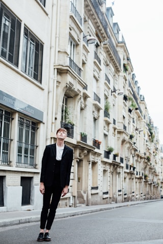 A person in a black suit and white shirt stands on an empty urban street with tall, elegant buildings lining the sidewalk. The architecture features classic European design, with balconies and large windows.