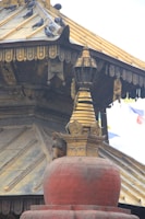 A monkey perches on a large, red, dome-shaped stupa structure with intricate golden detailing. Several pigeons are perched on the sloping roof above. The structure has ornate carvings and is adorned with religious or cultural symbols.