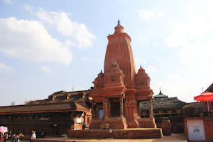 A traditional Hindu temple made from red brick, featuring intricate architectural details and a tall central spire. The courtyard setting includes other historic structures, with a mix of people milling around the area. The sky above is a clear blue with scattered clouds, adding to the scenic backdrop.