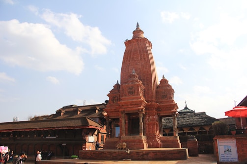 A traditional Hindu temple made from red brick, featuring intricate architectural details and a tall central spire. The courtyard setting includes other historic structures, with a mix of people milling around the area. The sky above is a clear blue with scattered clouds, adding to the scenic backdrop.