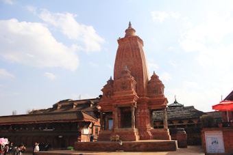 A traditional Hindu temple made from red brick, featuring intricate architectural details and a tall central spire. The courtyard setting includes other historic structures, with a mix of people milling around the area. The sky above is a clear blue with scattered clouds, adding to the scenic backdrop.