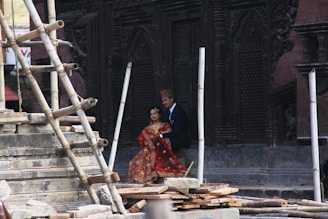 A couple dressed in traditional attire stands beside an intricately carved, historical structure with wooden scaffolding and stonework around them.