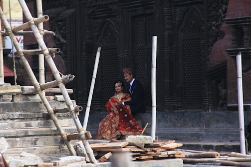 A couple dressed in traditional attire stands beside an intricately carved, historical structure with wooden scaffolding and stonework around them.
