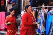 Two women dressed in traditional red attire are walking past a colorful street market. They are holding trays, possibly containing offerings. Behind them, various garments in a range of colors are displayed on hangers.