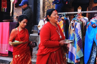 Two women dressed in traditional red attire are walking past a colorful street market. They are holding trays, possibly containing offerings. Behind them, various garments in a range of colors are displayed on hangers.