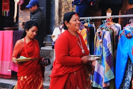 Two women dressed in traditional red attire are walking past a colorful street market. They are holding trays, possibly containing offerings. Behind them, various garments in a range of colors are displayed on hangers.