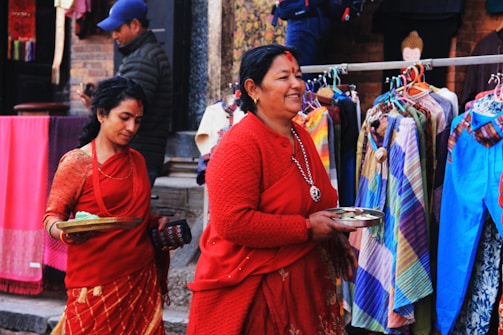 Two women dressed in traditional red attire are walking past a colorful street market. They are holding trays, possibly containing offerings. Behind them, various garments in a range of colors are displayed on hangers.