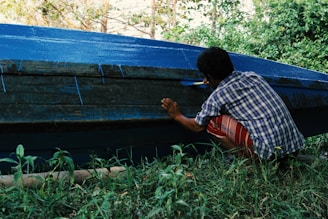 A person wearing a plaid shirt and striped shorts is crouched down, painting the side of a boat with blue paint. The boat is partially painted and is positioned on grassy ground, with trees visible in the background. The scene suggests an outdoor setting, possibly near a body of water.