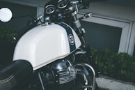 A close-up view of a motorcycle featuring a white gas tank with a logo, chrome accents, and a black leather seat. The handlebars include a rear-view mirror and control switches. The instrument cluster is visible with several gauges. The background includes green foliage and a concrete surface.