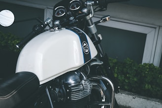 A close-up view of a motorcycle featuring a white gas tank with a logo, chrome accents, and a black leather seat. The handlebars include a rear-view mirror and control switches. The instrument cluster is visible with several gauges. The background includes green foliage and a concrete surface.