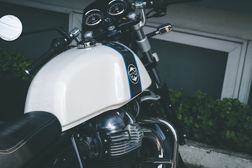 A close-up view of a motorcycle featuring a white gas tank with a logo, chrome accents, and a black leather seat. The handlebars include a rear-view mirror and control switches. The instrument cluster is visible with several gauges. The background includes green foliage and a concrete surface.