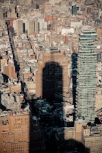 A dense urban landscape featuring a variety of buildings, including a distinctive modern skyscraper with a stacked design. The shadow of a tall building is cast prominently across the scene, adding a dramatic contrast to the tightly packed city blocks.