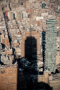 A dense urban landscape featuring a variety of buildings, including a distinctive modern skyscraper with a stacked design. The shadow of a tall building is cast prominently across the scene, adding a dramatic contrast to the tightly packed city blocks.