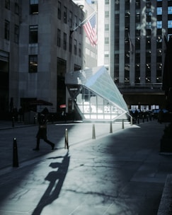 A city scene featuring a modern geometric glass structure and an American flag. The area is surrounded by tall urban buildings, casting long shadows on the pavement. A person is walking across the street, and the atmosphere is characterized by overcast lighting.