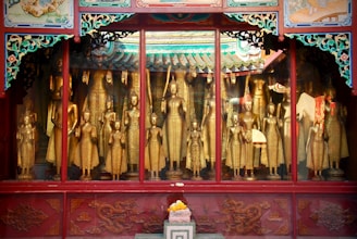 A collection of golden Buddha statues is displayed behind a glass window. Each statue varies in size and posture, surrounded by ornate red and green decorative frames. The intricate details in the carvings and vibrant patterns give a sense of historical and cultural richness.