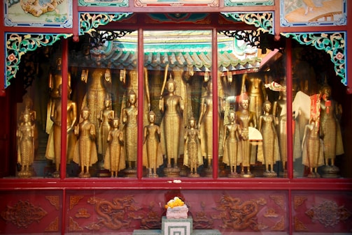 A collection of golden Buddha statues is displayed behind a glass window. Each statue varies in size and posture, surrounded by ornate red and green decorative frames. The intricate details in the carvings and vibrant patterns give a sense of historical and cultural richness.
