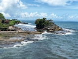 A scenic coastal location featuring a temple perched on a rocky outcrop, surrounded by blue ocean water. The area is bustling with tourists walking along the shore and enjoying the view. The coastline is lush with greenery, while waves crash against the rocks.