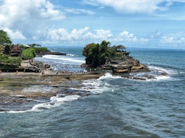 A scenic coastal location featuring a temple perched on a rocky outcrop, surrounded by blue ocean water. The area is bustling with tourists walking along the shore and enjoying the view. The coastline is lush with greenery, while waves crash against the rocks.