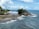 A scenic coastal location featuring a temple perched on a rocky outcrop, surrounded by blue ocean water. The area is bustling with tourists walking along the shore and enjoying the view. The coastline is lush with greenery, while waves crash against the rocks.
