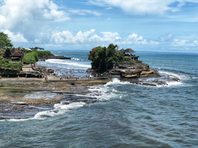 A scenic coastal location featuring a temple perched on a rocky outcrop, surrounded by blue ocean water. The area is bustling with tourists walking along the shore and enjoying the view. The coastline is lush with greenery, while waves crash against the rocks.