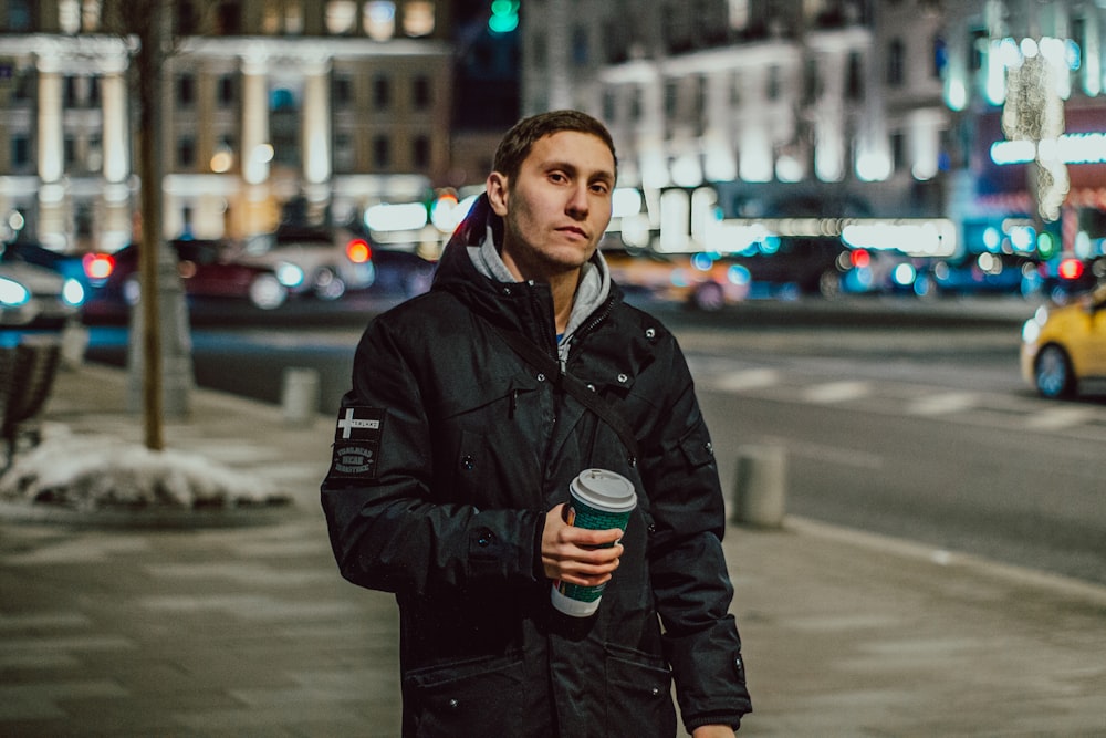 man in black jacket standing on sidewalk near busy road during night time