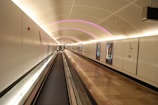A modern, illuminated walkway tunnel with a moving walkway. The tunnel is adorned with sleek white walls, some of which display posters. The ceiling features a series of soft, glowing purple lights that accentuate the curvature of the tunnel. Two individuals can be seen walking in the distance.