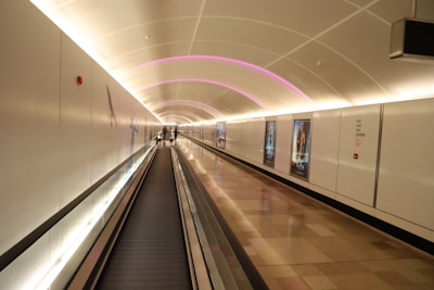 A modern, illuminated walkway tunnel with a moving walkway. The tunnel is adorned with sleek white walls, some of which display posters. The ceiling features a series of soft, glowing purple lights that accentuate the curvature of the tunnel. Two individuals can be seen walking in the distance.
