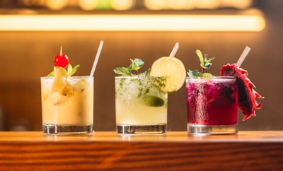Three colorful cocktails are lined up on a wooden bar counter. The first cocktail is a creamy yellow drink garnished with a red cherry and mint leaves. The second drink is light green with a lime wheel and mint sprig. The third cocktail is a vibrant magenta color with a slice of dragon fruit and mint.