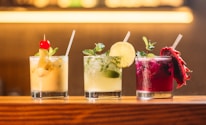 Three colorful cocktails are lined up on a wooden bar counter. The first cocktail is a creamy yellow drink garnished with a red cherry and mint leaves. The second drink is light green with a lime wheel and mint sprig. The third cocktail is a vibrant magenta color with a slice of dragon fruit and mint.