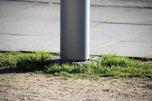 A vertical metal pole is fixed into a paved surface surrounded by patches of green grass and soil. The pole is cylindrical and rises from a metal base. The surrounding area features a mix of natural elements like grass and man-made materials like pavement.