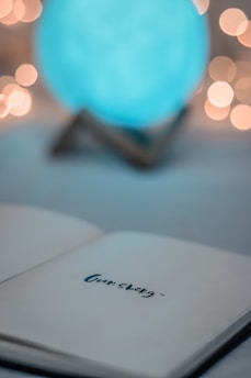 An open notebook displays the word 'Gursberg' written on one of its pages. In the background, a blurred, illuminated globe emits a soft blue glow, surrounded by warm bokeh lights.