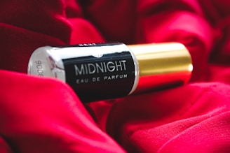 A bottle of 'Midnight Eau De Parfum' is resting on a rich, red fabric. The bottle features a sleek black and gold design, creating a luxurious and elegant aesthetic.