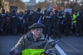 A person wearing a beret and glasses is in the foreground, possibly on a mobility device. In the background, a line of police officers stands facing a group of protesters during a demonstration. The atmosphere appears tense with a clear division between the officers and the crowd.