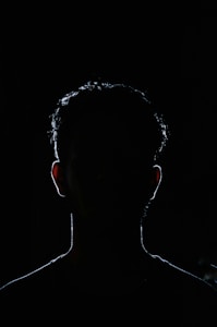 A dark silhouette of a person is visible against a black background. The outline of the head and shoulders is faintly illuminated, creating a mysterious and shadowy figure. Details of facial features are obscured by darkness, enhancing the enigmatic quality.