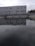 A large industrial building with multiple windows is situated behind a body of water, possibly part of a water treatment facility. A metal structure extends over the water, and trees are visible in the background under a cloudy sky.