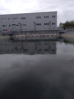 A large industrial building with multiple windows is situated behind a body of water, possibly part of a water treatment facility. A metal structure extends over the water, and trees are visible in the background under a cloudy sky.