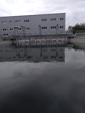 A large industrial building with multiple windows is situated behind a body of water, possibly part of a water treatment facility. A metal structure extends over the water, and trees are visible in the background under a cloudy sky.