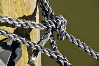 Several thick, black and white braided ropes are tightly knotted around a wooden post. The wood appears weathered with visible grain and some signs of wear. The ropes create a complex knot, displaying their texture and thickness against the smooth surface of the post.