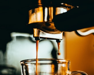 A coffee machine pouring freshly brewed espresso into a clear glass cup. The warm, golden lighting adds a cozy and inviting atmosphere, highlighting the dark, rich coffee being dispensed.