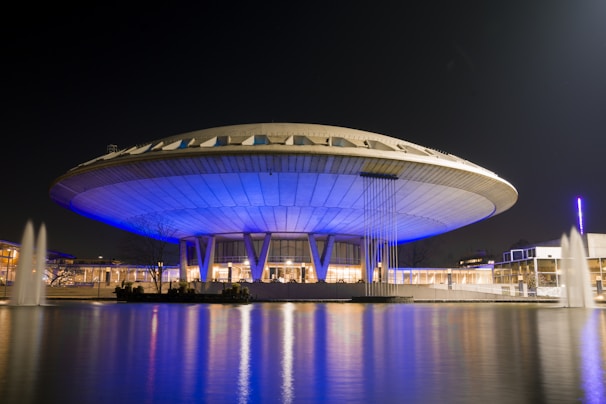 A UFO-shaped building is illuminated with blue lighting at night, creating a futuristic appearance. The structure is surrounded by water, reflecting the lights and adding to the serene ambience.
