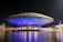 A UFO-shaped building is illuminated with blue lighting at night, creating a futuristic appearance. The structure is surrounded by water, reflecting the lights and adding to the serene ambience.