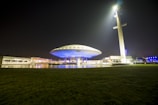 A futuristic building resembling a UFO is illuminated at night, with a large spotlight beaming from a nearby tall tower. The structure features a circular design with bright blue lights, surrounded by a grassy area and reflections in a water feature.