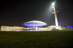 A futuristic building resembling a UFO is illuminated at night, with a large spotlight beaming from a nearby tall tower. The structure features a circular design with bright blue lights, surrounded by a grassy area and reflections in a water feature.